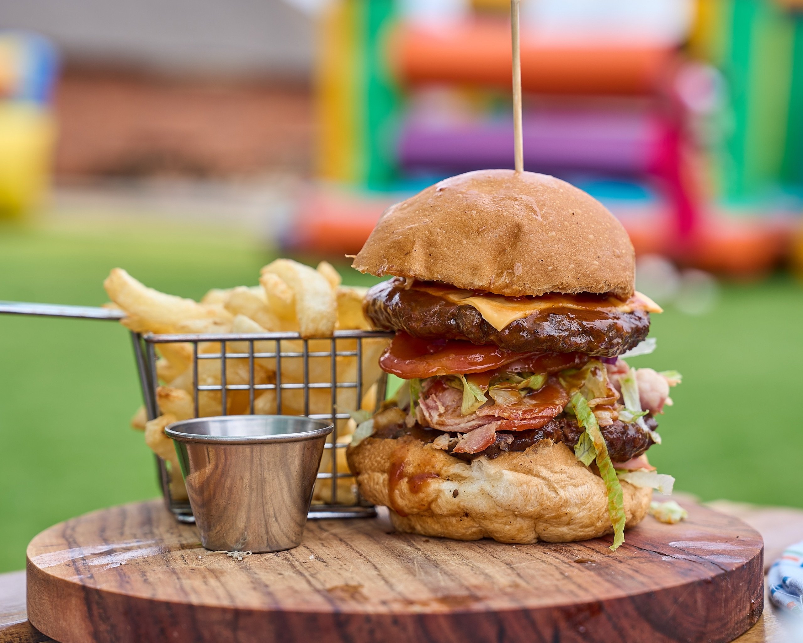 Stacked burger with bacon and a basket of fries, bouncy castle behind